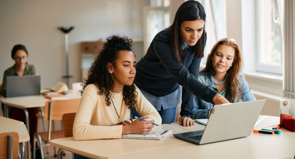 A woman stands between two female students pointing to a laptop screen.