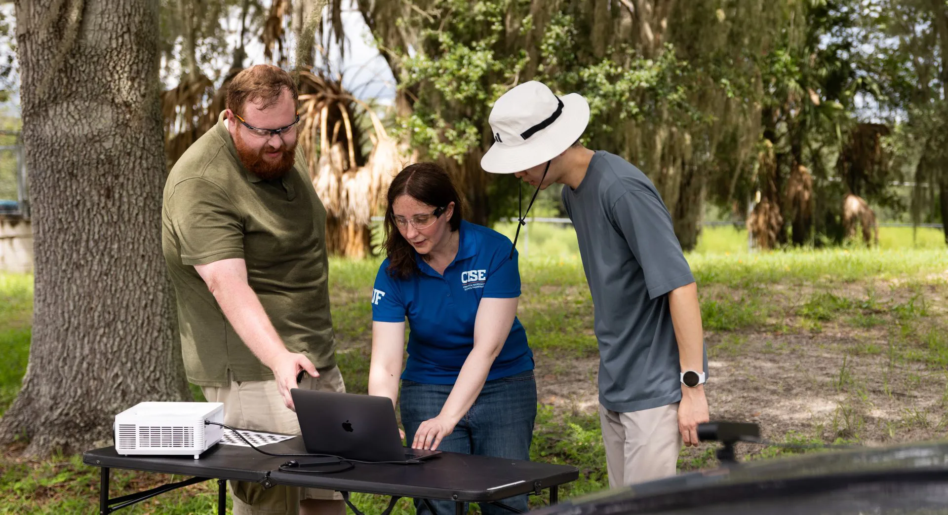 Three researchers stand and look at a computer in a green outdoors area
