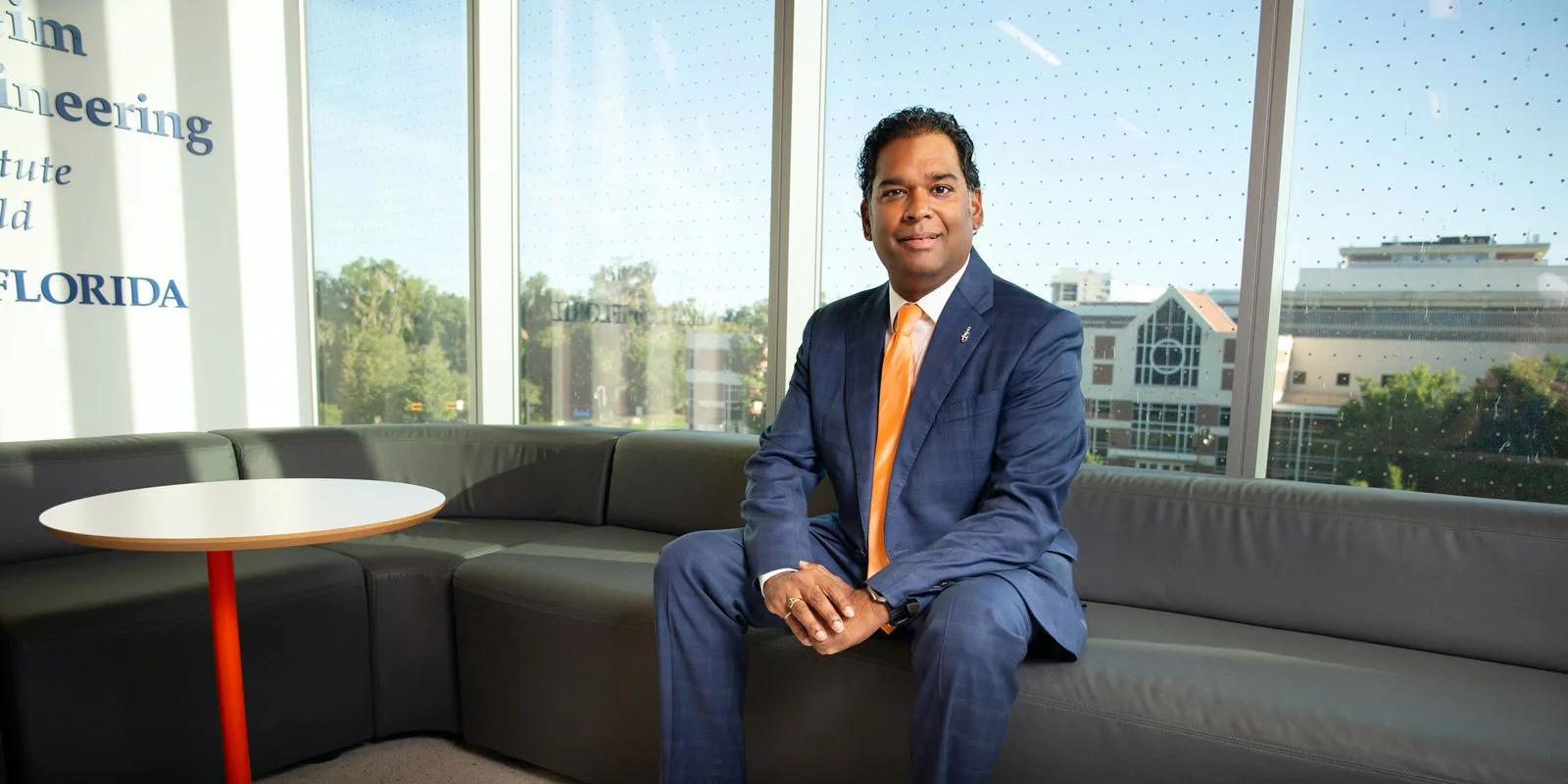 A man in a blue suit and orange tie sits on a gray sectional sofa in a bright room with large windows overlooking buildings and trees.